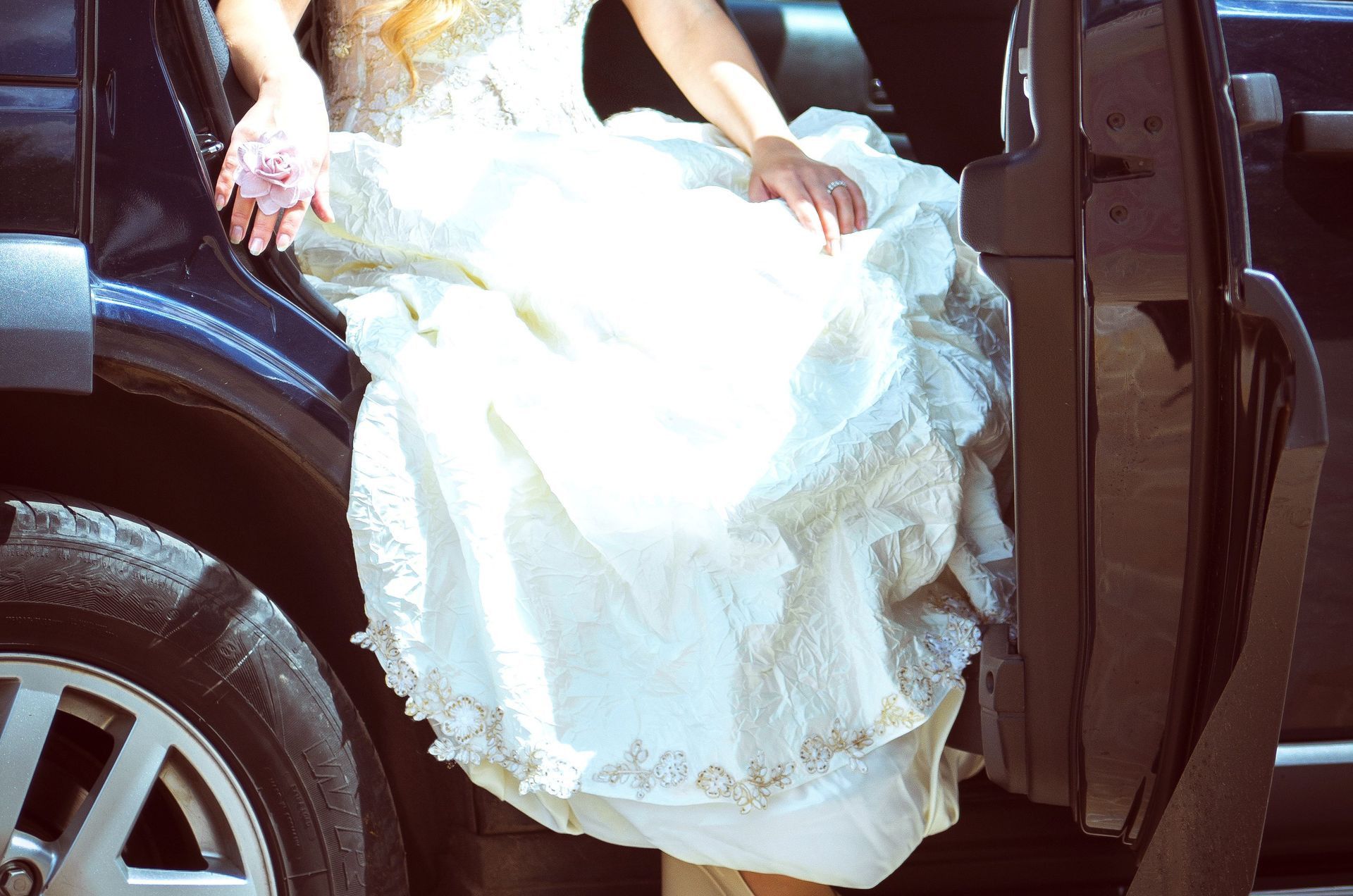 Elegant bride in a wedding dress gracefully sits in a car on a sunny day, ready for the ceremony.